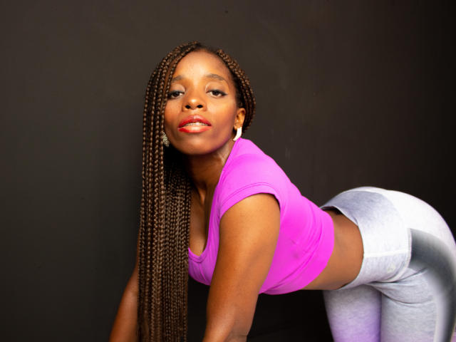 SandyChaudeX with long braids, pink crop top, and gray butt-hugging pants, bending over, showing her ass, looking at the camera with a slight smile.