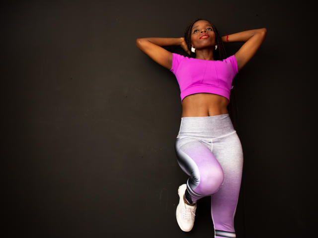 SandyChaudeX with dark skin, braids, wearing a pink crop top, grey and purple gradient leggings, white sneakers, arms behind head, right leg bent, confident, sexy.