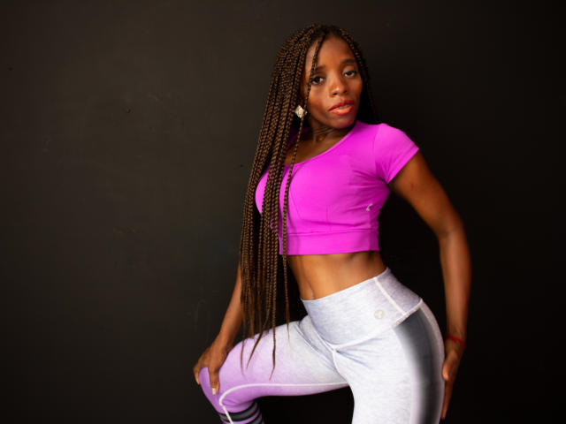 SandyChaudeX with dark skin, long braids, pink crop top, showing abs, white pants, one knee up, looking at camera, black background, subtle tattoo on right hand.