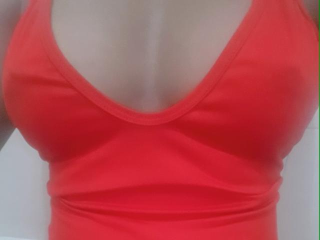 Karhinna's red tank top is pulled tight, showing her sizable, rounded boobs with a deep V-neck. Her skin is light, and the image focuses on her chest.
