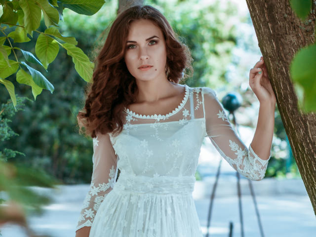 AnissaLogan with wavy brown hair, wearing a white lace dress with floral patterns, standing by a tree, holding a branch, green leaves in the foreground.