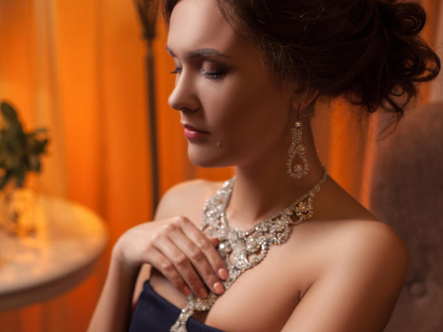 AnissaLogan has her hand on her tits, wearing a glittery diamond necklace and matching earrings, with dark hair in an updo, in a warm, orange-lit room.