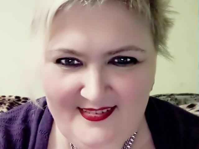LanaNighty tiene el cabello rubio corto, lápiz labial rojo y una camisa morada, sonriendo con su pequeña nariz linda y delineador de ojos negro. Lleva un collar de plata..