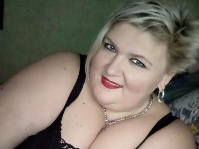 LanaNighty, una donna bionda di taglia grande con i capelli corti, rossetto rosso, una maglia di pizzo nero e una collana d'argento, sorride alla fotocamera, mostrando il suo seno prosperoso..