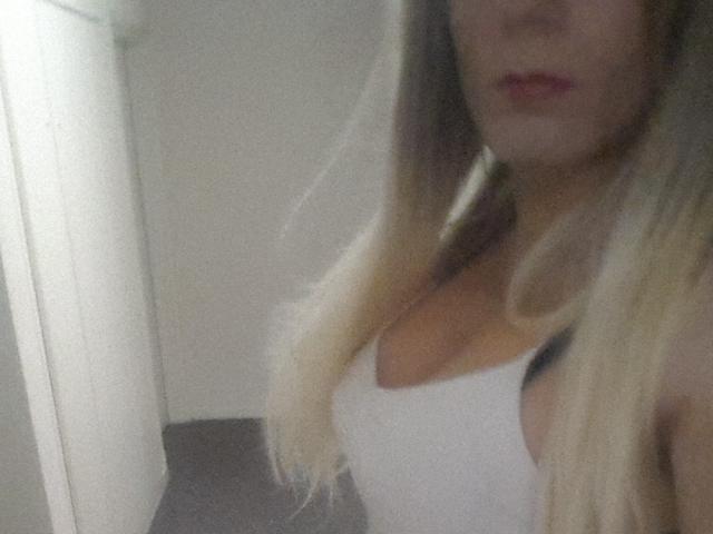 Alessiacia mit blonden Haaren, einem weißen Tanktop und freiliegendem Dekolleté, die ihre Brüste zeigt, leicht unscharf, in einem schwach beleuchteten Raum. Ihre Lippen sind leicht geöffnet..