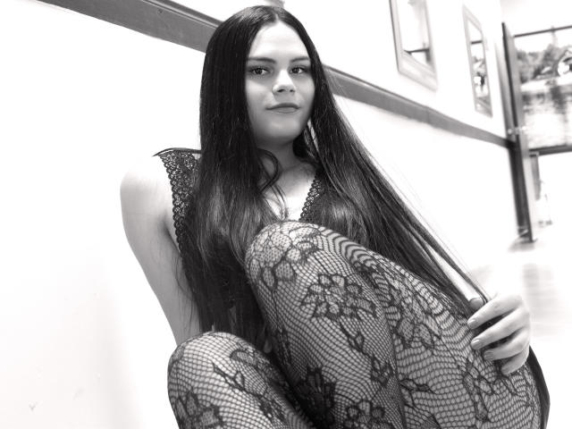 JassmineTS with long dark hair, lace-patterned fishnet stockings, and lace top, squatting, looking at camera, black-and-white, confident, sexy, legs crossed, slight smile.