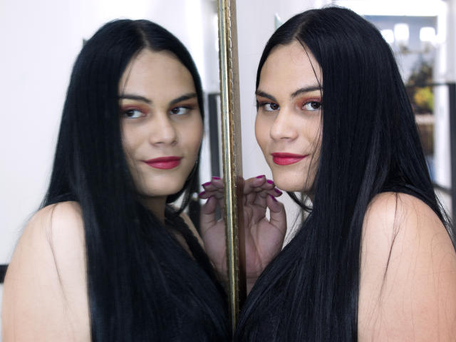JassmineTS, avec ses longs cheveux noirs, son rouge à lèvres rouge et ses ongles violets, se tient devant un miroir, montrant son reflet avec un sourire discret, tout en tenant ses cheveux..