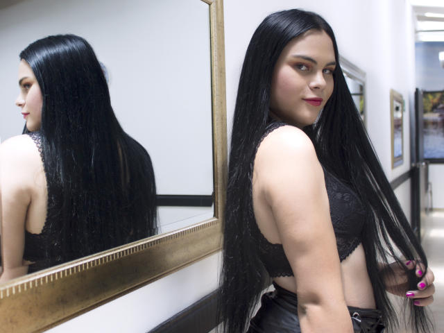 JassmineTS, con lunghi capelli neri, indossa un reggiseno di pizzo nero, è in piedi di fronte a uno specchio, mostrando la schiena e sorridendo, e il suo riflesso è visibile nello specchio. Il suo seno è visibile..