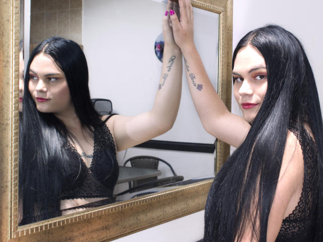 JassmineTS with long black hair, wearing a black lace bra, reflects in a mirror. She's looking at her reflection, showing tattoos on her arm. Her tits are small.