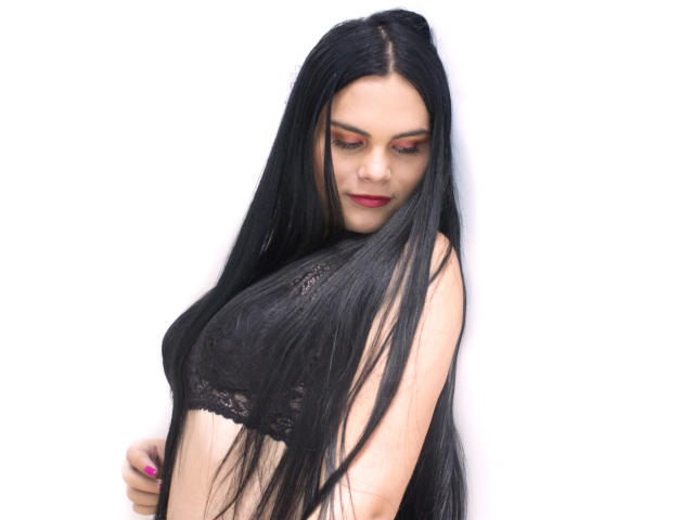 JassmineTS mit langen, schwarzen Haaren, trägt einen schwarzen Spitzen-BH, hat roten Lippenstift und rosafarbene Nägel, blickt leicht lächelnd nach unten und zeigt ihre Brüste. Weißer Hintergrund..
