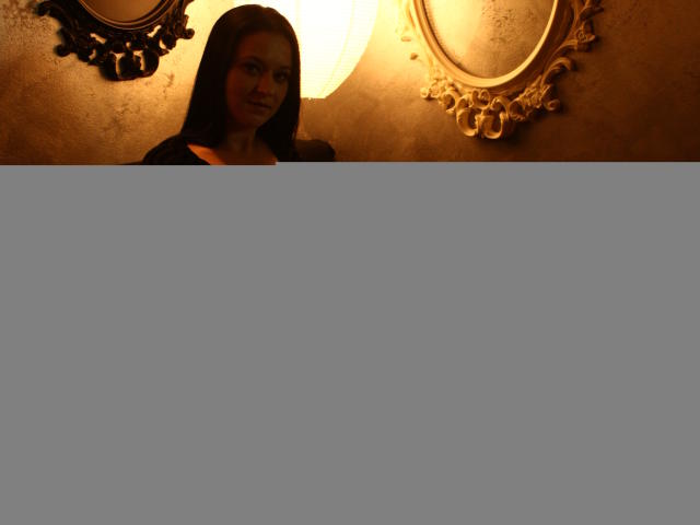 SensualCrissa with dark brown hair, sitting on a black couch, wearing a low-cut black dress, exposing her tits, under warm, yellow lighting, with ornate mirrors in the background.