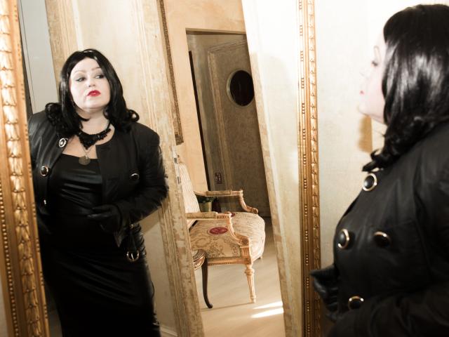 KaryQueen with black hair, red lipstick, and black leather dress stands in front of a mirror, reflecting her full-ass and busty figure. She wears black gloves and a necklace.