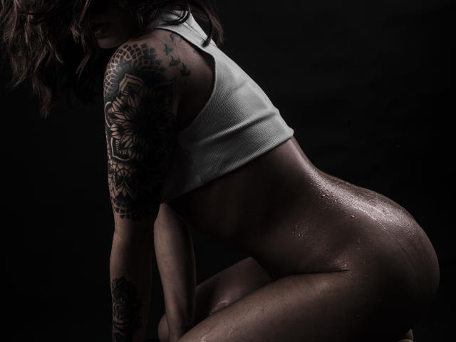 KassiKassou, con la pelle umida, un top corto bianco e tatuaggi, è accovacciata, mostrando il suo sedere sudatore e un lato del seno. L'immagine è scura, suggestiva e sensuale. Il tatuaggio sul suo braccio è dettagliato e ben definito..