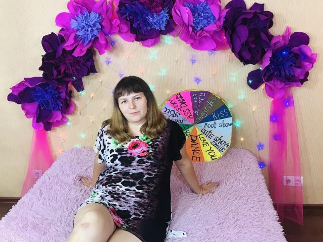AliceInks, with brown hair and bangs, wearing a leopard print dress with a pink flower, sits on a pink fuzzy bed, surrounded by purple and blue paper flowers.