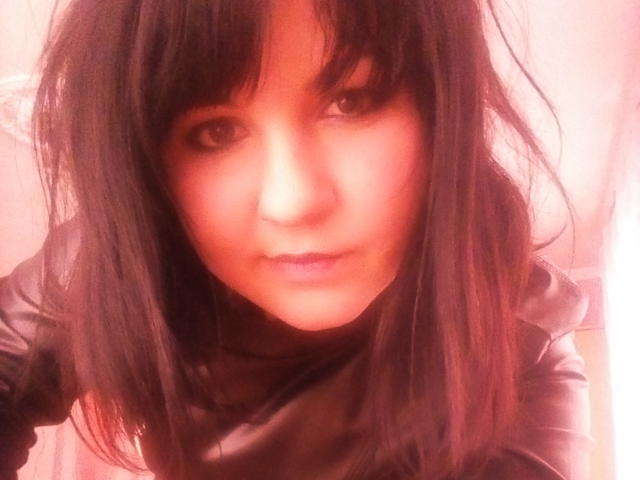 AnnaBloon, avec ses cheveux noirs, regardant vers le haut avec un sourire subtil, portant une chemise noire, éclairage rosé, mise au point douce, cheveux légèrement en désordre, et une expression mignonne, légèrement séduisante..