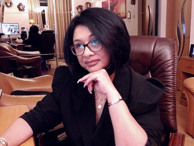 ClassybutNaughty with black bob, glasses, and black top, her hand resting on her chin, a bracelet on her wrist, sitting in a leather chair, smiling slightly.