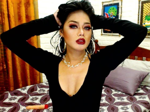 Un uomo con un'erezione, le mani tra i capelli neri, indossa una maglia nera a V profondo, rossetto rosso, una collana di perle e grandi orecchini a cerchio, in piedi di fronte a un letto con lenzuola a fantasia..