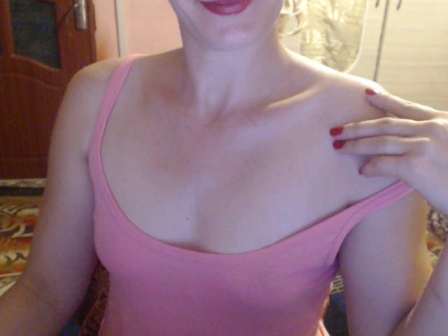 SweetLoves mit heller Haut, einem pinken Tanktop, roten Nägeln und rotem Lippenstift, die ihre kleinen Brüste, das leicht sichtbare Schlüsselbein und ihre rechte Hand auf der Schulter zeigt..