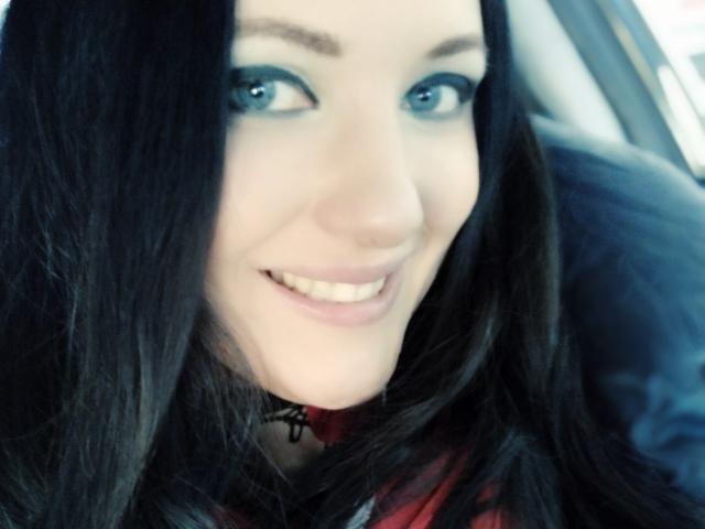 Le visage de SweetttAngel, avec ses yeux bleus, ses cheveux noirs et lisses, et son sourire blanc éclatant. Elle porte un haut rouge et noir et est assise dans une voiture..