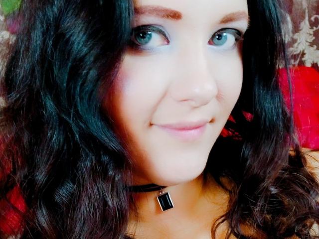 SweetttAngel has blue eyes, wavy dark hair, a slight smile, and wears a black choker with a square gem. Her skin is fair and she looks cute.