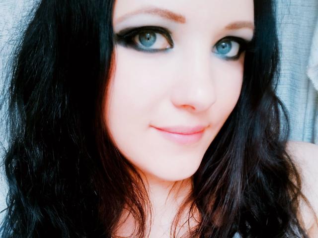 SweetttAngel has blue eyes, black eyeliner, and a slight smile with wavy dark hair, a pale face, and pink lips. Her tits are not visible in this shot.