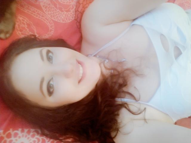 SweetttAngel with curly brown hair, white tank top, and a necklace, lying on a red bed, smiling at the camera, showing her tits and a cute face.