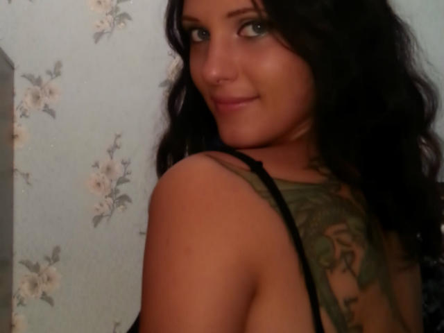 SweetttAngel has wavy black hair, light skin, and a tattoo on her back. She's smiling, showing her tits, with a black strap on her shoulder.