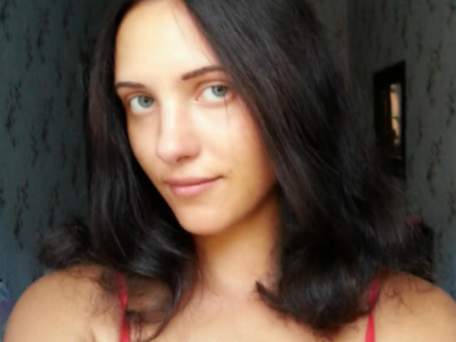 SweetttAngel has fair skin, green eyes, and dark brown wavy hair. She's wearing a red spaghetti-strap top and has a subtle smile on her face.