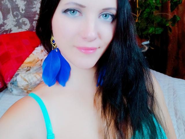 SweetttAngel with blue feather earrings, light blue eyes, pink lips, black hair, green tank top, subtle smile, pale skin, and a small nose piercing. Boobs visible, upper body.