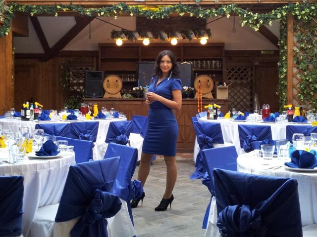 SweetttAngel, mulher curvilínea em um vestido azul justo, segurando uma bebida, em pé em frente a mesas com decorações azuis e brancas, usando saltos pretos, sorrindo para a câmera..