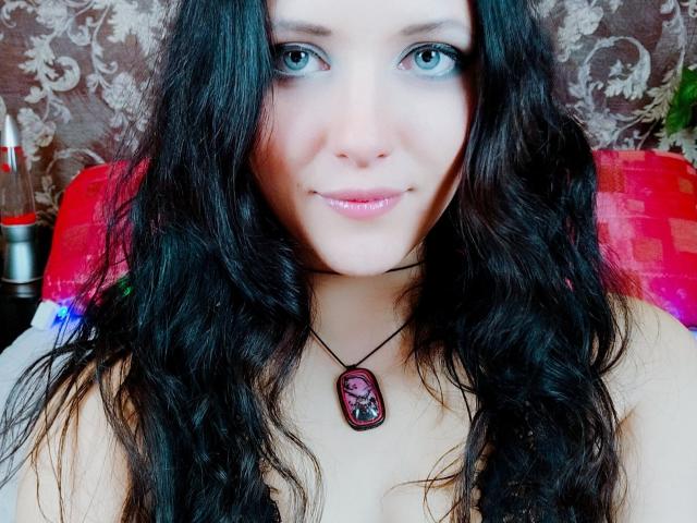 SweetttAngel com longos cabelos pretos ondulados, olhos azuis e lábios rosados, usando um colar preto com uma pedra roxa, olhando para a câmera com um sorriso discreto..