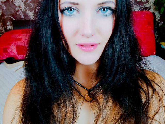 SweetttAngel hat leuchtend blaue Augen, langes schwarzes Haar, rosafarbene Lippen und trägt eine schwarze Halskette mit einem braunen Anhänger. Ihre nackten Schultern sind sichtbar..