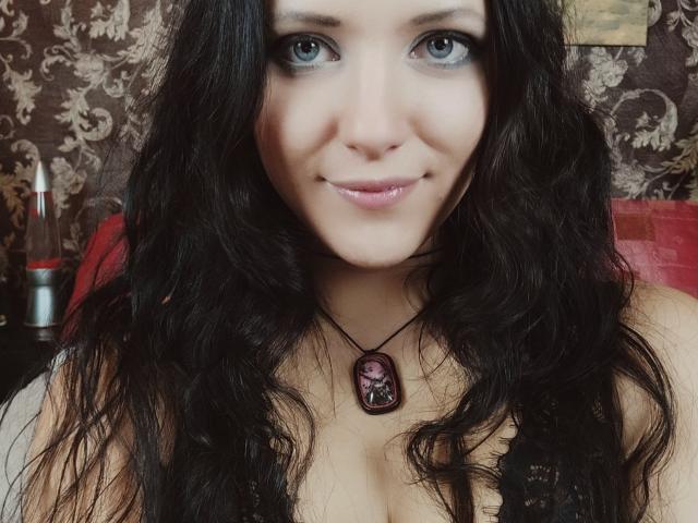 SweetttAngel with wavy black hair, blue eyes, and a slight smile, wearing a black lace top, and a pendant with a purple gem, showing her tits.