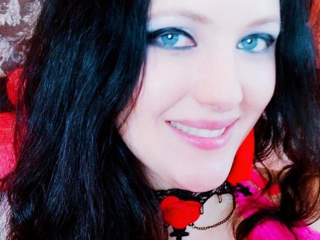 SweetttAngel with blue eyes, black wavy hair, pink lipstick, smiling, wearing a black lace choker with red rose accents, looking directly at the camera.