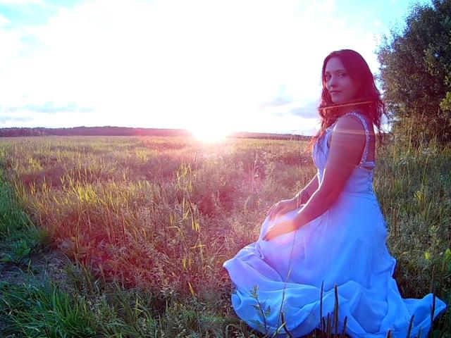 SweetttAngel with wavy dark hair, wearing a white dress, sitting in a grassy field at sunset, her ass slightly visible, sun glowing behind her, gentle light on her face.