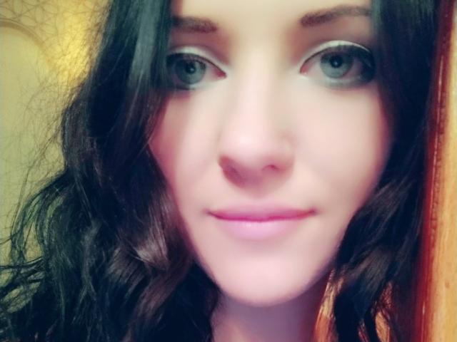 A selfie of SweetttAngel with blue eyes, wavy dark brown hair, and pink lips, showing a subtle smile. Her nose is small and her skin is fair.