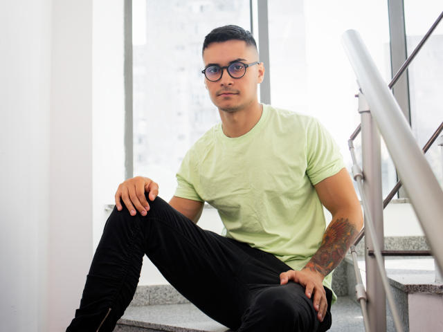 MattMalone sitting on stairs, wearing a green tee, black pants, glasses, and boots, with tattooed arm, looking serious, hands on knees, metal railing to the right.