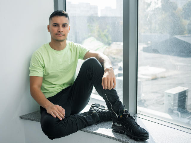 MattMalone, gespierd, bruine huid, kort zwart haar, groene T-shirt, zwarte broek, zwarte laarzen, zittend op een vensterbank, serieuze uitdrukking, zonlicht, stedelijke achtergrond, rechterbeen gekruist, linkerarm rustend..