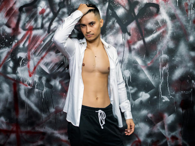 MattMalone, shirtless with a white button-up, black pants, and shaved head, hand in hair, standing against graffiti. He has a slim build, light skin, and a serious look.