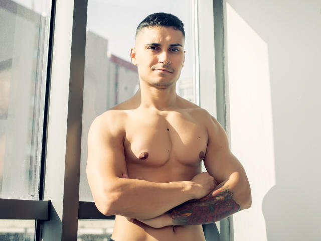 MattMalone, ohne Hemd und mit verschränkten Armen, hat einen muskulösen Körperbau, ein Tattoo am rechten Unterarm, kurze, dunkle Haare und ein leichtes Lächeln. Seine Brustwarzen und sein Penis sind sichtbar..