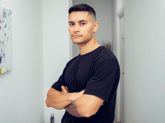MattMalone, em pé com os braços cruzados, vestindo uma camiseta preta, cabelo preto curto, tatuagem no antebraço direito, expressão neutra, pele clara, em um quarto com paredes brancas..