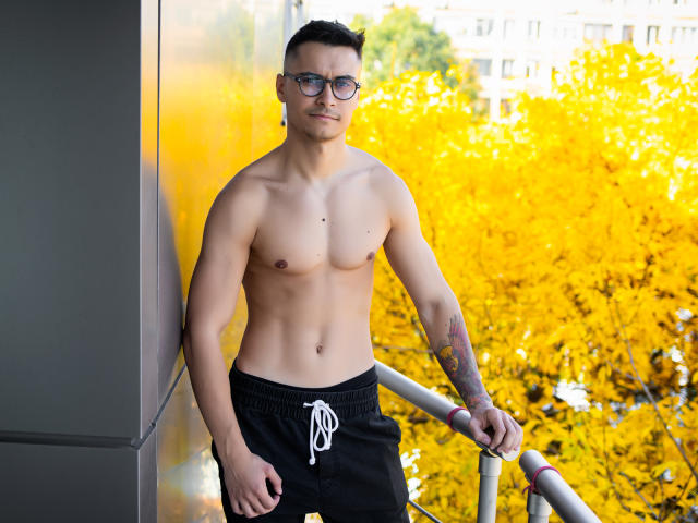 MattMalone está sem camisa, vestindo shorts pretos com um cordão branco, em pé perto de um corrimão, usando óculos e tem uma tatuagem no antebraço direito. Árvores amarelas brilhantes ao fundo..