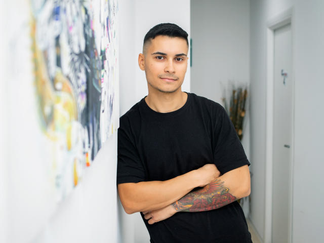 MattMalone with short black hair, black t-shirt, arms crossed, tattoo on right forearm, standing in white hallway, smiling slightly, brown eyes, light skin, medium build, cute.