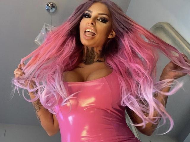 BarbaraBae con capelli che sfumano dal rosa al viola, la lingua fuori, indossa una top rosa in lattice lucido, mettendo in mostra il suo seno prosperoso e i tatuaggi, e tiene i suoi lunghi capelli..