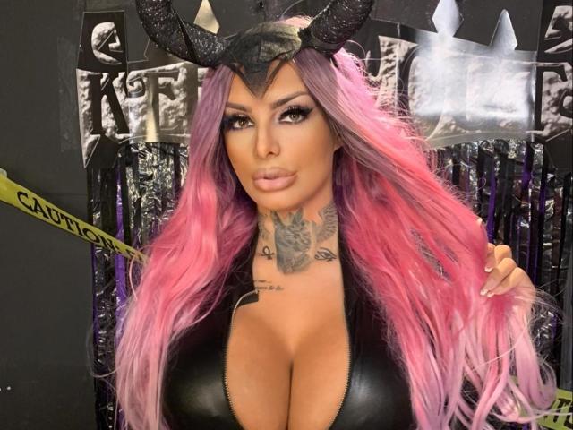 BarbaraBae mit rosa Haaren, einem schwarzen Kopfschmuck in Form von Hörnern und einem Ausschnitt in einem schwarzen Top, der ein Tattoo an ihrem Hals zeigt. Ihre Brüste sind groß und ihr Make-up ist auffällig..