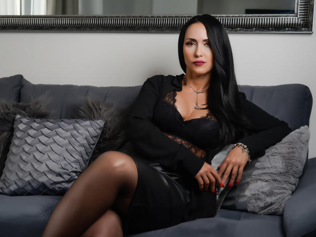 MissVonTease with long black hair, wearing a black lace bra, black dress, and sheer pantyhose, sitting on a gray couch with textured pillows, looking confident.