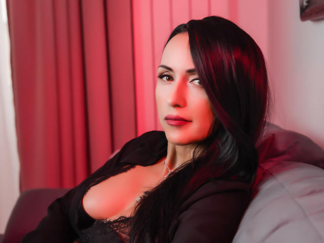 MissVonTease has dark, shiny hair, red lips, and is looking to the side with a slight smile. She's wearing a black top that reveals her big, shiny tits.