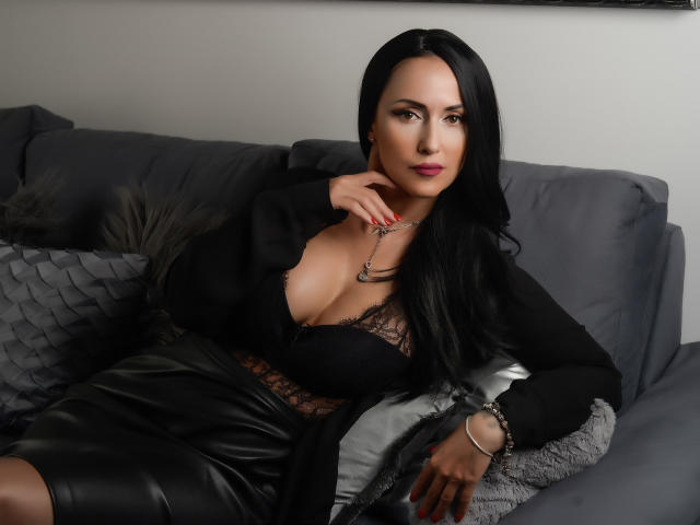 MissVonTease, con i capelli neri, indossa una maglia di pizzo nero e una gonna in pelle, seduta su un divano grigio, che mette in mostra il suo seno prosperoso e il suo décolleté sexy, con una collana..