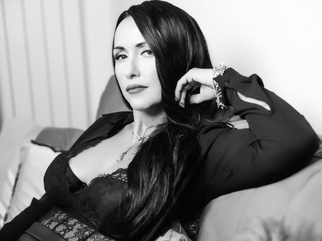 MissVonTease with long dark hair, wearing a black lace bra and open shirt, sitting on a couch, looking to the side, right hand in hair, black and white.