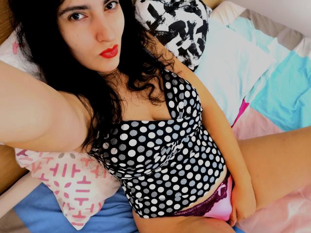 AddictedToUTS has black hair, red lipstick, and wears a polka dot top, pink lace panties, showing a hand on their pussy, lying on a bed.